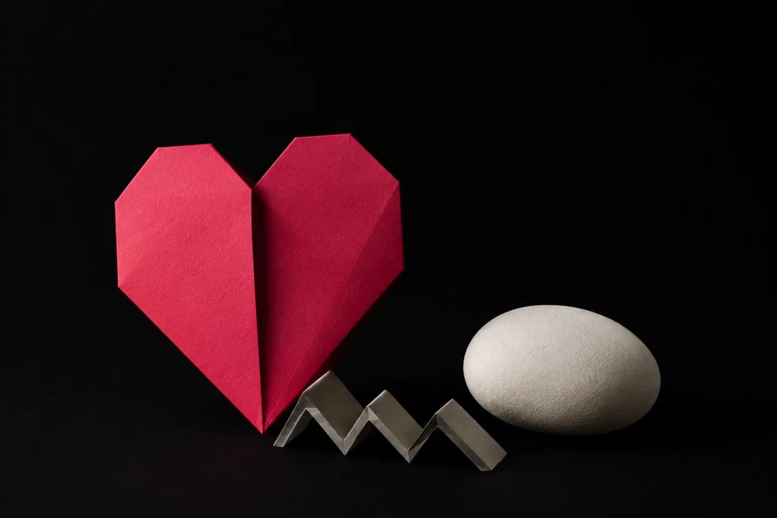 A folded paper heart beside a small zigzag and a smooth pebble.